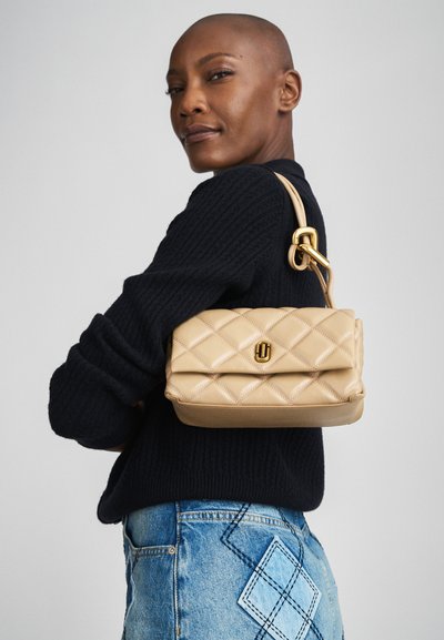 Beige quilted handbag with a smooth texture, a gold hardware clasp, and a short strap, worn against a dark sweater and denim.
