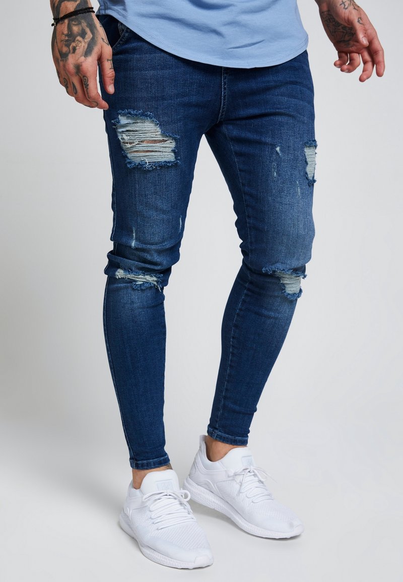Blå denim skinny jeans med rivna detaljer, med en figurnära design och en lätt sliten textur, kombinerade med vita träningsskor.