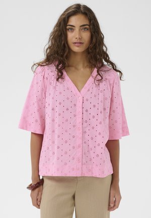 Femme aux cheveux bouclés portant un chemisier rose à boutons avec œillets et manches courtes, ainsi qu'un pantalon beige, debout devant un fond uni.