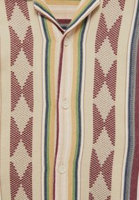 Camicia a maniche corte con bottoni, realizzata in un tessuto testurizzato con strisce verticali multicolori e motivi a diamante in rosso, crema e blu.