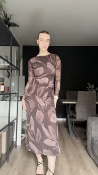 Vestido de manga longa em castanho escuro com estampado floral claro. Design ajustado com detalhes franzidos; mangas transparentes e a bainha até ao tornozelo. Sandálias pretas com tiras.