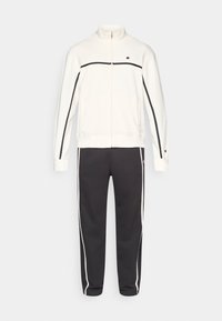 Λευκό τζάκετ με φερμουάρ και μαύρες λεπτομέρειες, με ψηλό γιακά, συνδυασμένο με σκουρόχρωμο γκρι παντελόνι με στενή γραμμή και άσπρες ρίγες στο πλάι.