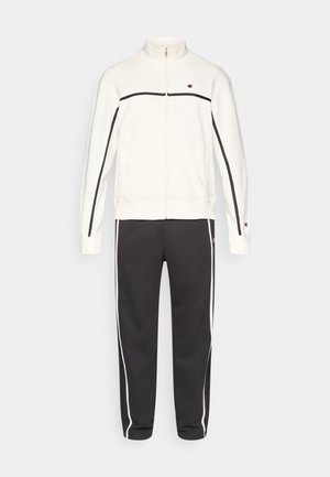 Veste de survêtement blanche à fermeture éclair avec des accents noirs et un col montant, associée à un pantalon gris foncé fuselé avec des bandes blanches sur les côtés.