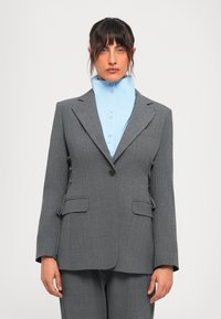Woman wearing a gray suit with a single button and a high-collared light blue shirt, standing against a plain white background.