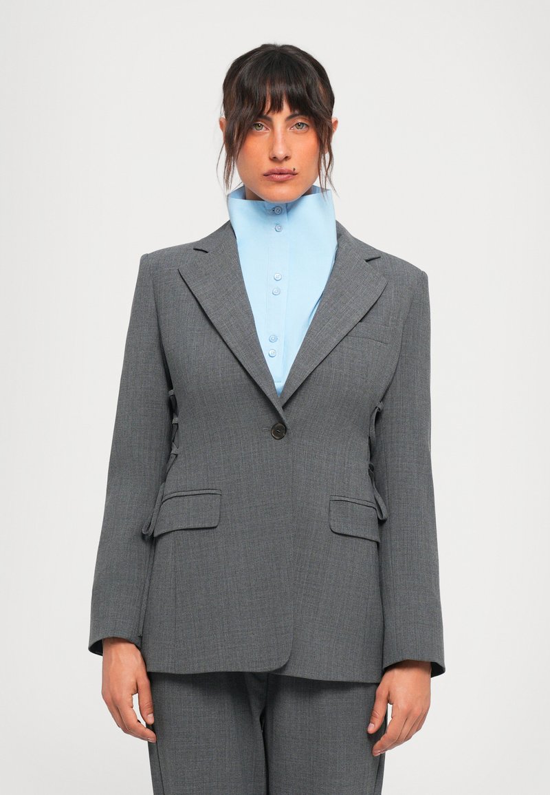 Woman wearing a gray suit with a single button and a high-collared light blue shirt, standing against a plain white background.