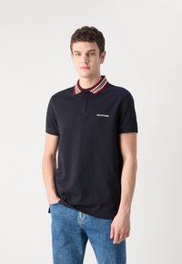 Ναυτικό polo μπλουζάκι με ριγωτό γιακά σε κόκκινο, λευκό και πράσινο χρώμα, που διαθέτει ένα μικρό κεντητό λογότυπο στο στήθος. Φοριέται με μπλε τζιν.