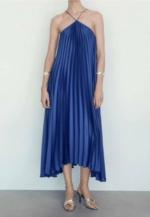 Une robe plissée longueur cheville en tissu bleu riche, avec des bretelles fines et un ourlet évasé, accompagnée de talons argentés à lanières.