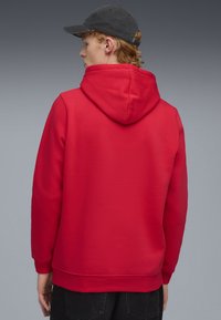 Rode hoodie met een trekkoordcapuchon, lange mouwen en geribbelde manchetten. Glad textuur, afgewerkt met een rechte zoom. Model draagt een zwarte pet.