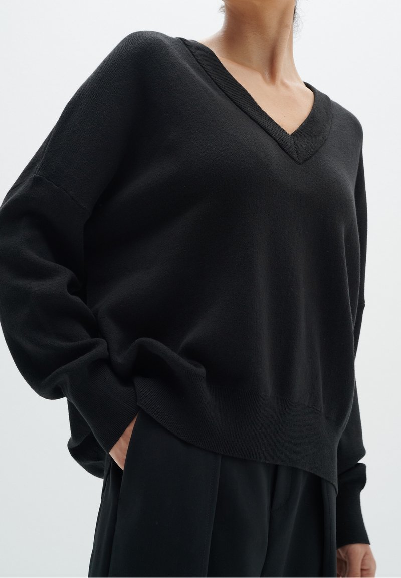 Personne portant un pull noir ample à col en V et un pantalon noir, une main dans la poche, sur un fond clair uni.