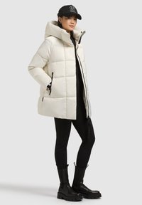 Biela prešívaná bunda s kapucňou, predným zipsom, bočnými vreckami a čiernymi akcentmi. Nosená cez čierne legíny a masívne čierne topánky.