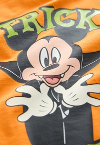 Orange T-shirt med en tegneseriefigur, der er afbildet med et sort kappe, hvide handsker og et legende udtryk mod en orange baggrund.