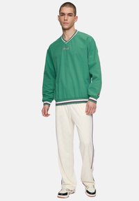Groene sweatshirt met een v-hals en contrasterende afwerking, gecombineerd met crèmekleurige broek met een subtiel patroon en donkere zijstrepen.