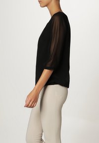 Femme en blouse noire à manches transparentes et pantalon beige clair ajusté, debout de profil devant un fond uni.
