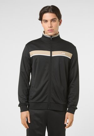 Młody mężczyzna w czarnej sportowej kurtce na zamek marki FILA z beżowymi i białymi paskami na wysokości klatki piersiowej, stojący na neutralnym tle.