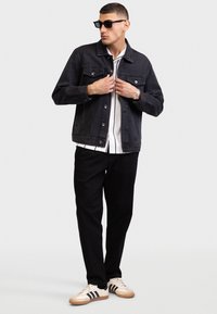 Dark denim jacket with a collar, front pockets, and button closures, worn over a white shirt and black pants, paired with casual sneakers.