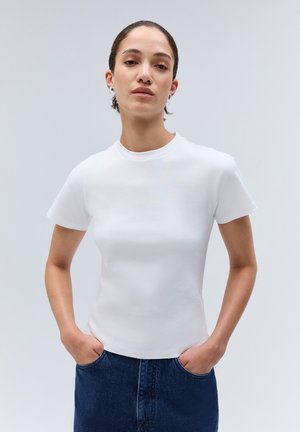 Femme aux cheveux courts et foncés portant une chemise blanche à manches courtes et un jean bleu, debout les mains dans les poches devant un fond uni.