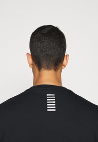 T-shirt nero con scollo rotondo e una striscia riflettente verticale sulla parte alta della schiena. Tessuto morbido con una texture liscia.