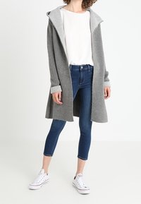 Grå lång cardigan med huva, mjuk textur, öppen front och uppvikt ärm. Bärs över en vit topp och kombineras med blåjeans och vita sneakers.