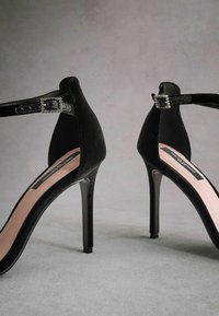 Scarpe con tacco alto in velluto nero con cinturino alla caviglia e fibbia decorativa. Presentano un design elegante e una punta affusolata, con una soletta rosa chiaro.