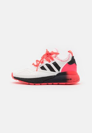 Basket de sport blanche avec des bandes noires, des lacets corail vifs et des accents corail et noirs sur la semelle, vue de côté sur fond blanc.