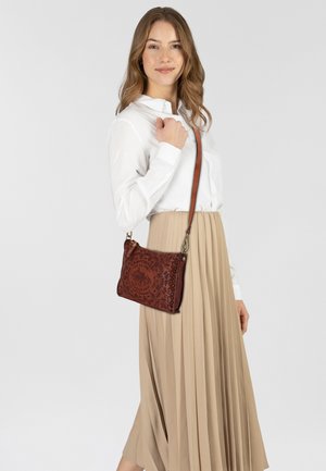 Borsa a tracolla in pelle marrone con design circolare a rilievo, superficie texturizzata e tracolla regolabile, indossata da una modella con camicia bianca e gonna beige.