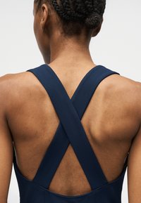 Άτομο με κοτσιδάκια που φοράει μανίκιαν μπλε ρούχο με φαρδιές σταυρωτές τιράντες στην πλάτη, μπροστά σε μονόχρωμο φόντο.