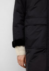 Hand som bär en krämfärgad tröja med svarta pälskanter, vilande bredvid en svart vadderad jacka med en stor klaffficka mot en neutral bakgrund.