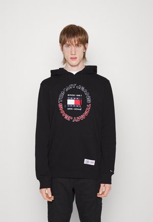 Felpa nera con cappuccio con una grafica circolare centrale che presenta il testo "Tommy Jeans" e un design a blocchi colorati. Include una tasca frontale e un'etichetta con il logo.