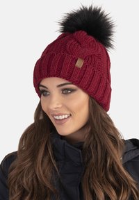 Gorro de punto burdeos con un patrón texturizado, que presenta un pompón de piel sintética negra en la parte superior y un pequeño etiqueta de cuero como acento.