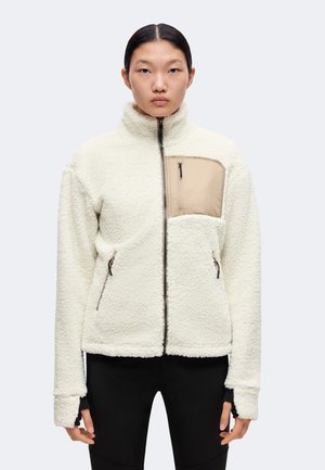 Vrouw met stijl zwart haar die een witte fleecejas draagt met een beige borstzak met rits en zwarte broek, staand tegen een effen achtergrond.