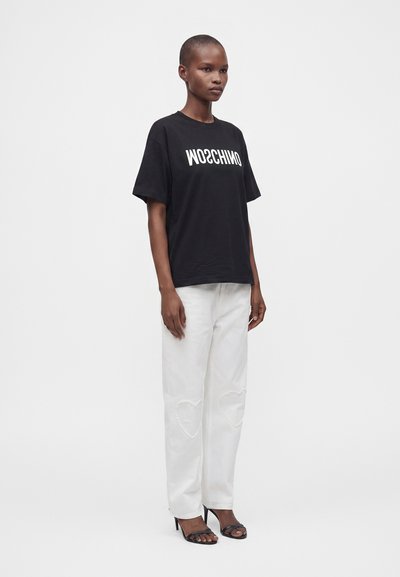 Άτομο όρθιο που φοράει μαύρο t-shirt Moschino, λευκό παντελόνι με σχέδια καρδιών στα γόνατα και μαύρα ανοιχτά παπούτσια σε απλό φόντο.