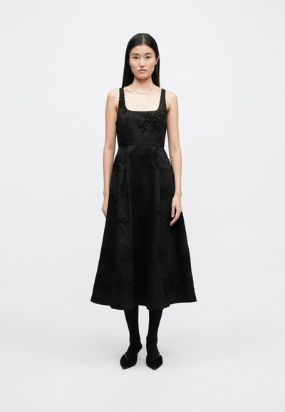 Μαύρο φόρεμα από σατέν με ρυθμιζόμενες τιράντες, διακοσμημένο με λεπτομέρειες από χάντρες και λουλούδια, σε ρέουσα A-line σιλουέτα και μήκος μέχρι το γόνατο.