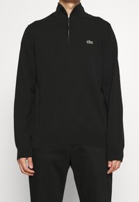 Svart kvartdraget tröja med ribbade ärmslut och nederkant, med en liten grön Lacoste-logotyp på vänster bröst. Mjuk tygtextur.