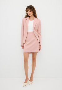 Blazer rosa pálido con un solo botón, falda a juego y blusa blanca; la modelo lleva mules en tono crema con punta; tejido suave, corte ajustado.