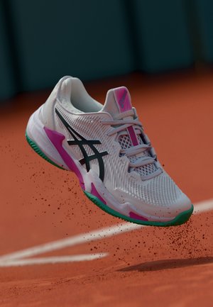 Białe sneakersy sportowe z czarnymi akcentami, różowymi i zielonymi detalami, z siateczkową teksturą górną i wyprofilowaną podeszwą, wydające się unosić nad glinianą powierzchnią.