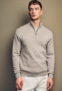Svijetlosivi pulover s teksturom i polu-zip ovratnikom, rebrastim manžetama i pojasom. Nosi se preko jednostavne sive košulje, uparen s bijelim hlačama.