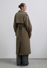 Langer, olivgrüner, gesteppter Mantel mit tailliertem Gürtel, übergroßen Ärmeln und doppelreihiger Button-Design, das ein geteiltes Rückenteil für Bewegungsfreiheit bietet.