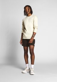 Beige genser med lange ermer, svarte shorts, hvite treningssko og hvite sokker. Modellen står mot en enkel lys bakgrunn.