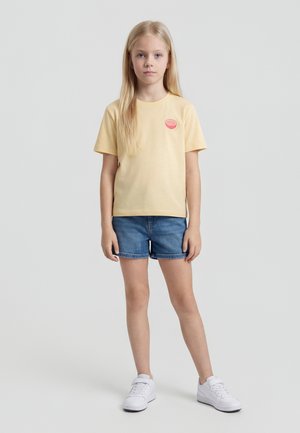 Fille blonde debout face à l'avant, portant un t-shirt jaune pâle avec un motif de fruits, un short en jean et des baskets blanches sur un fond gris uni.