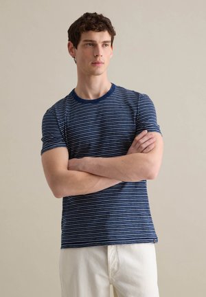 T-shirt blu navy a maniche corte con righe bianche orizzontali, colletto tondo e una texture morbida. Il modello sta in piedi con le braccia conserte, indossando pantaloni beige.