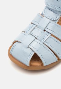 Light blue patent leather sandal with a woven design, open toe, and brown sole. Features smooth texture and contrasting stitching details.