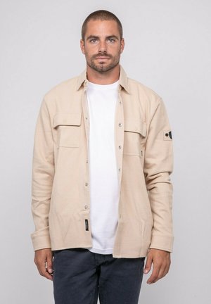 Homme aux cheveux courts et barbe, portant une chemise beige boutonnée par-dessus un t-shirt blanc et un pantalon foncé, debout devant un fond clair uni.