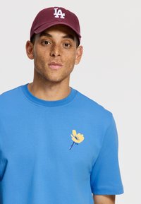 Camiseta de algodón azul con un gráfico de flor amarilla en el pecho. La gorra color berenjena tiene el logotipo "LA" bordado en blanco.