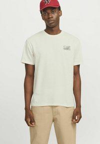 T-shirt di cotone crema con maniche corte, scollo rotondo e una piccola stampa grafica sul petto; abbinato a pantaloni beige.