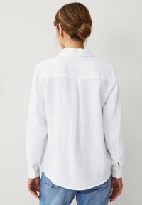 Camicia bianca di lino con maniche lunghe, colletto e polsini con bottone. Design con piega posteriore notevole e orlo leggermente curvo. Vestibilità casual.