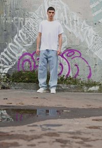Weißes Baumwoll-T-Shirt, hellblaue lockere Denim-Jeans und weiße Sneaker. Der Hintergrund zeigt eine strukturierte Wand mit lila Graffiti und grünem Moos.