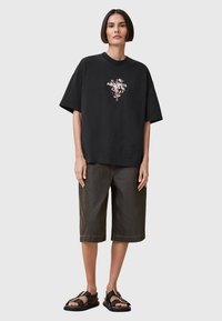 T-shirt oversize nera in cotone con grafica floreale e testo, abbinata a pantaloni corti in denim verde scuro e sandali neri con dettagli metallici.