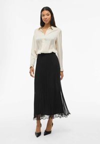 Satin crèmekleurige blouse met knoopdetails, gecombineerd met een zwarte geplooide midirok met doorschijnende accenten en een golvende zoom. Zwarte hakken maken de look compleet.