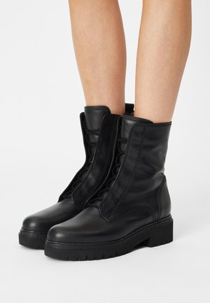 Sorte læderstøvler til kamp, der går op til midt på læggen, med tykke såler, båret af en person, der står på en hvid baggrund.