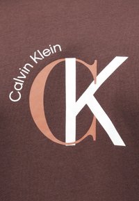 Hnedá bavlnená tkanina s veľkým grafickým motívom, na ktorom je napísané "Calvin Klein" v bielej farbe a "CK" v bielej a svetlo oranžovej farbe. Jednoduchý, odvážny dizajn.